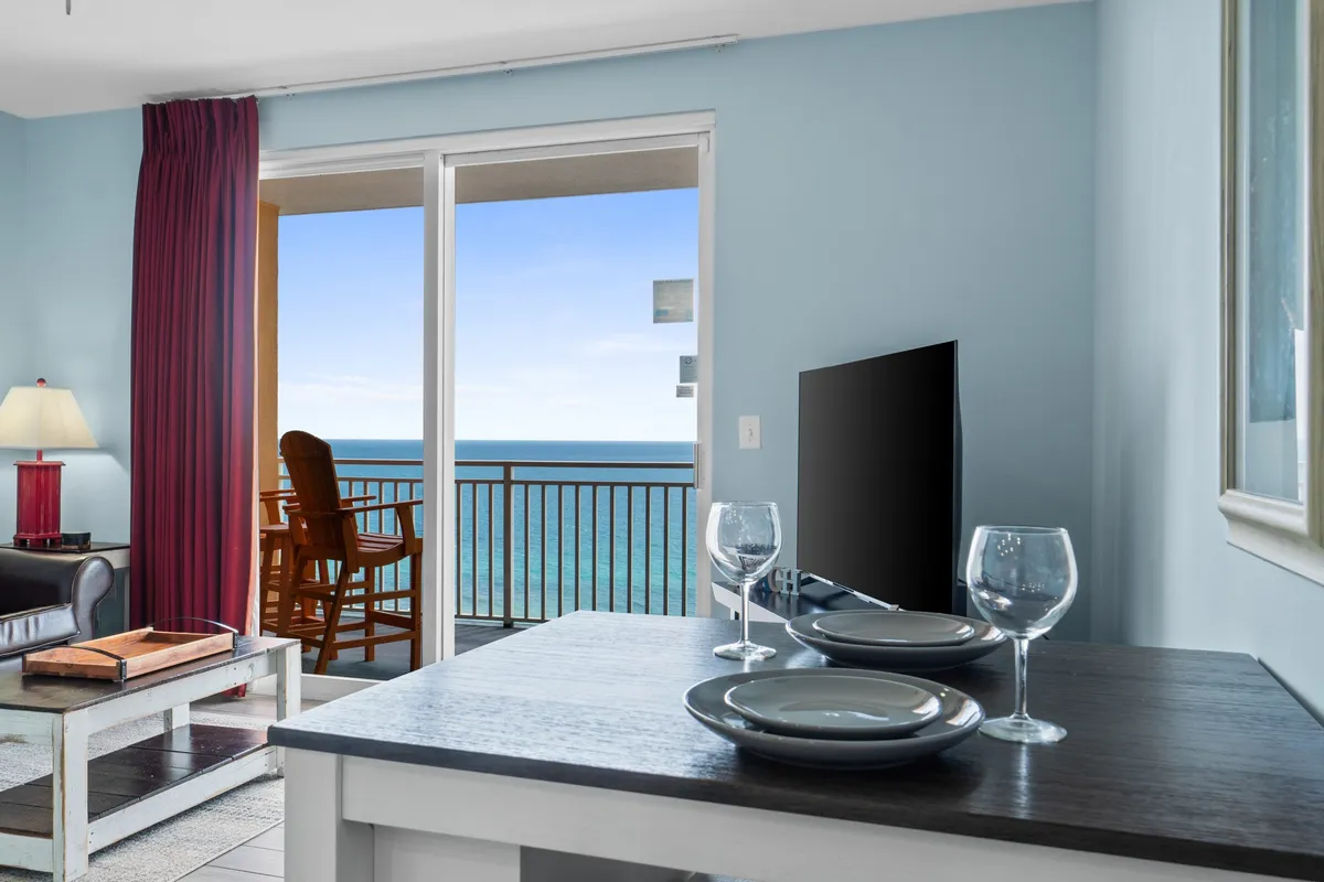 A dining area set with plates and glasses sits beside large sliding glass doors that open directly onto a private balcony. Through the doors, the ocean is visible, along with a comfortable chair on the balcony, suggesting opportunities for dining with a view, entertaining, or enjoying the breeze.