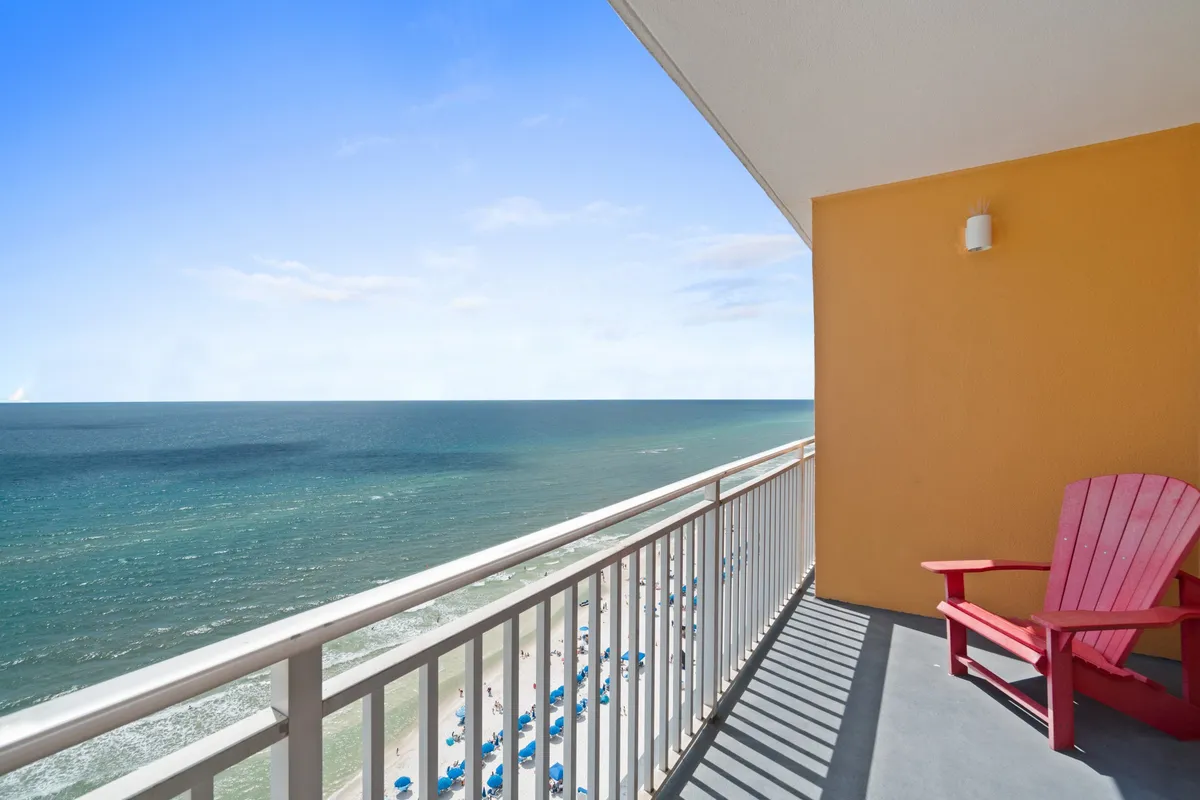A serene balcony scene overlooking the ocean, featuring a comfortable red chair and beach umbrellas. The tranquil blue waters of the Gulf stretch out to the horizon, capturing the sense of privacy and relaxation that a romantic or solo retreat promises.