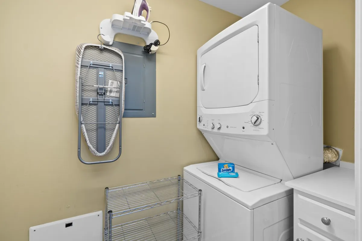 A practical laundry area with a washing machine, dryer, ironing board, and organized shelving unit for laundry supplies. The space is clean and efficiently arranged, emphasizing the convenience of in-unit amenities.