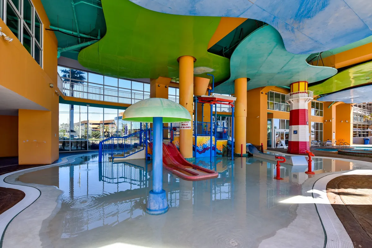 A vibrant indoor water park area featuring colorful water slides, a mushroom fountain, and interactive play structures, all under a cheerful, dynamic ceiling. Children are seen playing and splashing in the water, while families gather and relax poolside.