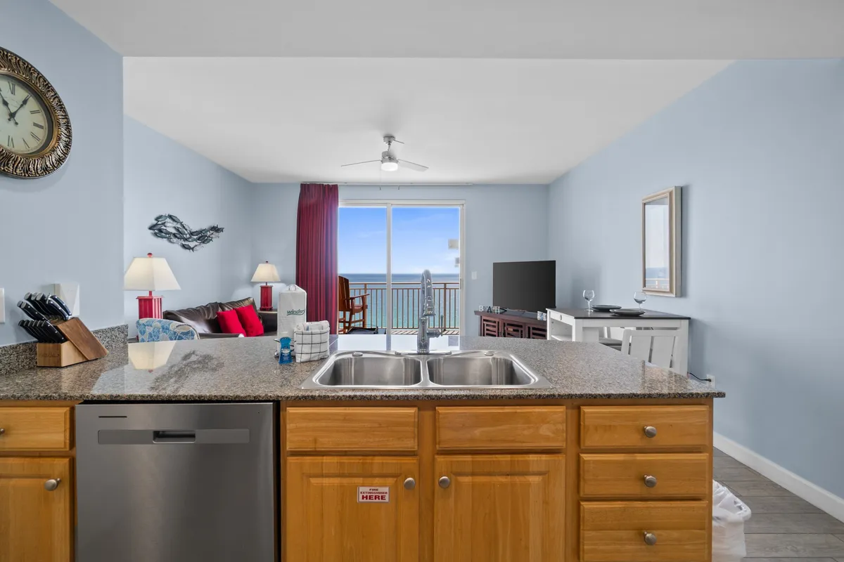 A view from the kitchen sink across a granite countertop and into a sunlit living area, with a window offering a glimpse of the ocean. The open-plan design keeps the chef connected to the rest of the group and frames meal prep as a shared experience.