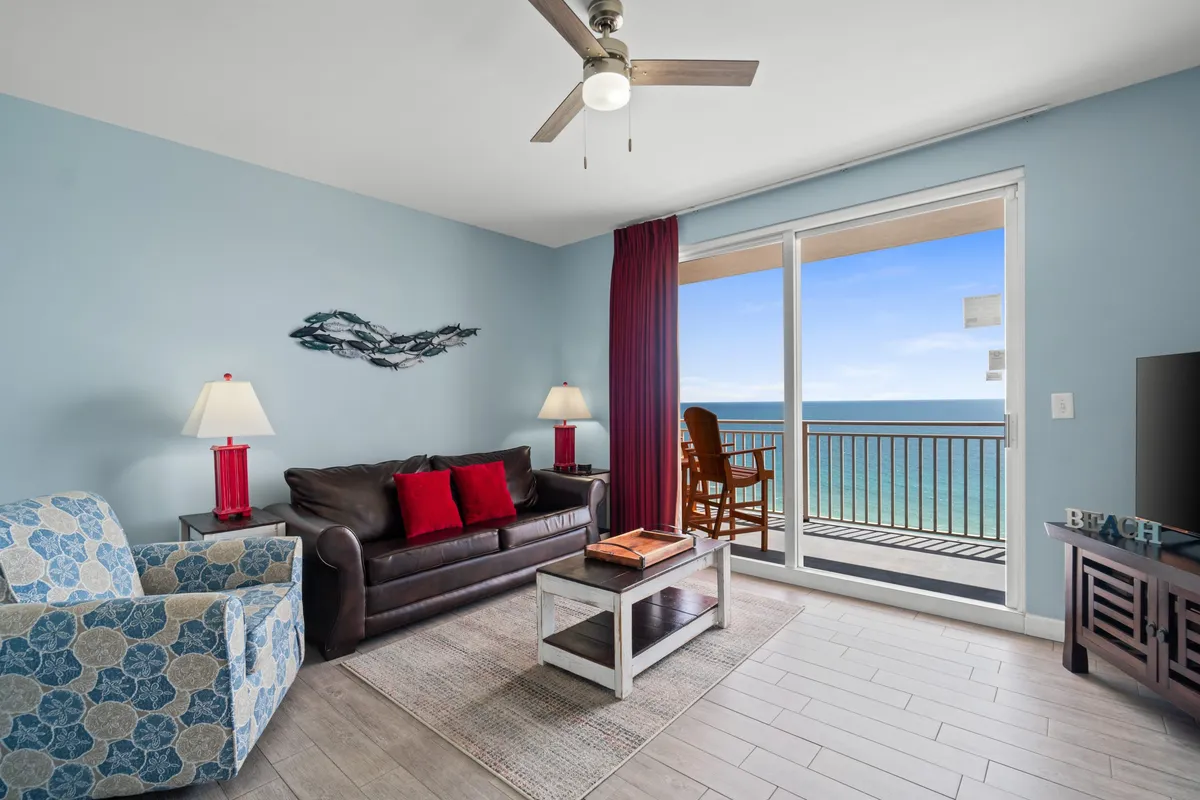 The image features a spacious living area with a dark leather sofa adorned with vibrant red accent pillows, a blue patterned armchair, and a floor-to-ceiling sliding glass door revealing breathtaking ocean views. The balcony outside is furnished with a wooden chair overlooking the Gulf, while the interior is decorated in a tropical coastal style, exuding comfort and warmth.