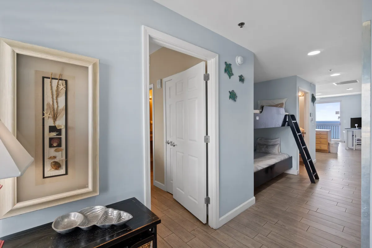 A family-friendly sleeping nook with bunk beds and a ladder, an open door leading to a bathroom, wall art with sea shells and starfish, and a view toward the living area with balcony and ocean view in the distance.