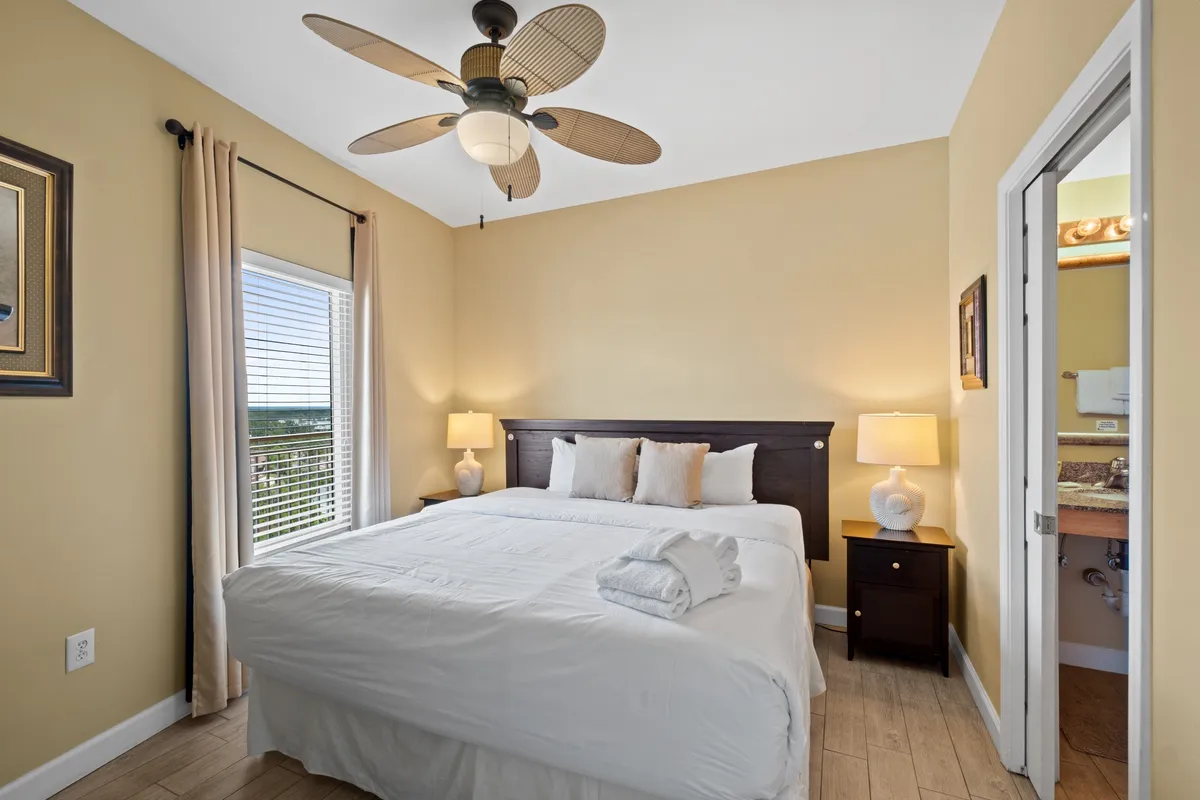 A spacious master bedroom featuring a king-size bed dressed in crisp white linens, folded towels placed at the foot, a window with beige curtains letting in soft daylight, a ceiling fan with leaf-shaped blades, and a doorway leading to a private bathroom.