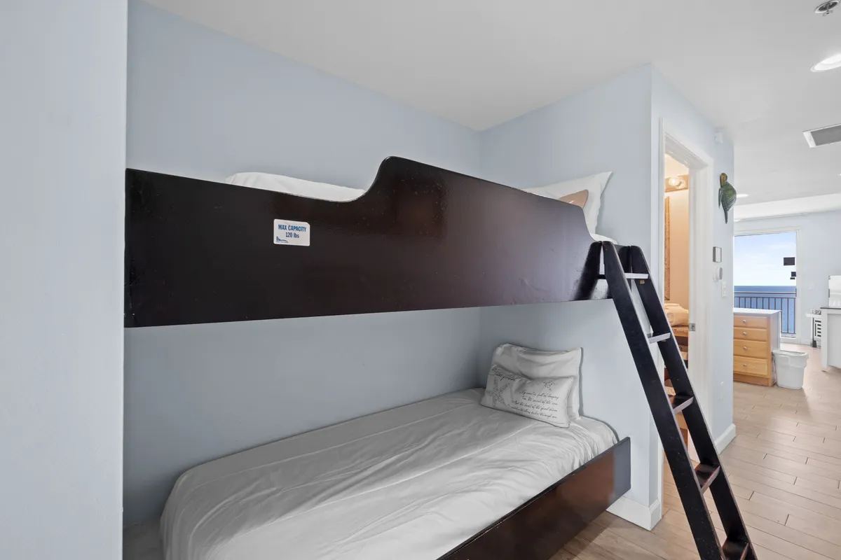 A cozy alcove with twin-over-twin bunk beds in a dark wood frame, a narrow ladder for easy access to the upper bunk, a hallway leading to bathroom and living areas, and a glimpse into the bright main living space with balcony access.
