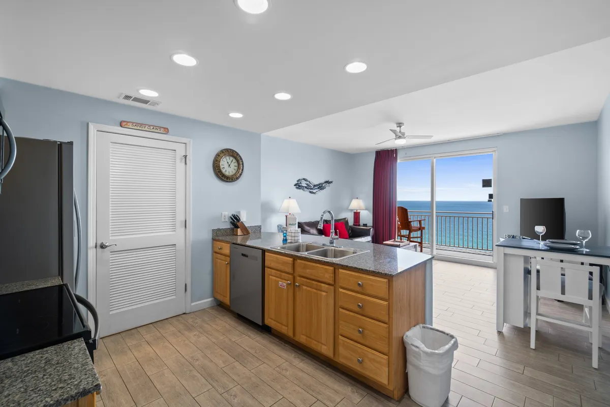 A bright and airy living space is highlighted by a red sofa, granite kitchen island, and expansive floor-to-ceiling windows opening to a private balcony with seating. The ocean view is a central feature, while the room’s decor combines modern and tropical elements, creating an inviting and lively atmosphere.