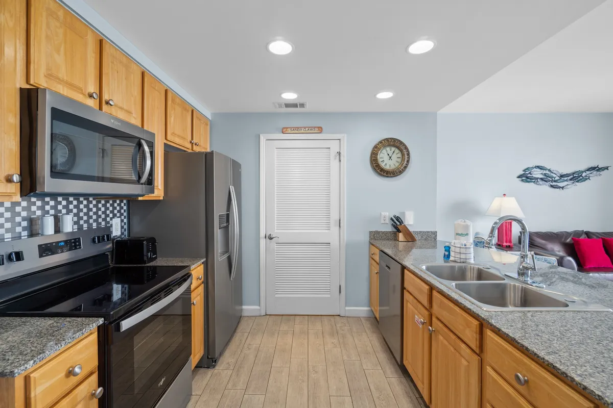 A bright, inviting kitchen with stainless steel refrigerator and oven, granite countertops, wooden cabinets, and a central kitchen island with a sink. The space is well-lit and organized for easy meal prep and family-friendly use.