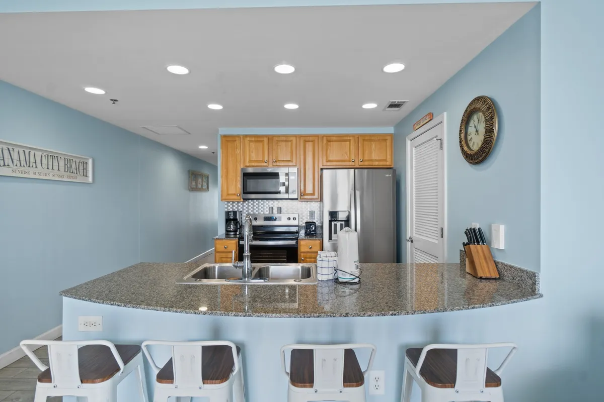 This image features a modern kitchen with granite countertops, a double sink, stainless steel appliances including a refrigerator and stove, wooden cabinetry, and bar seating. The blue painted walls and decorative clock add a touch of coastal charm. The bar stools at the counter invite casual dining and socializing within the kitchen space.