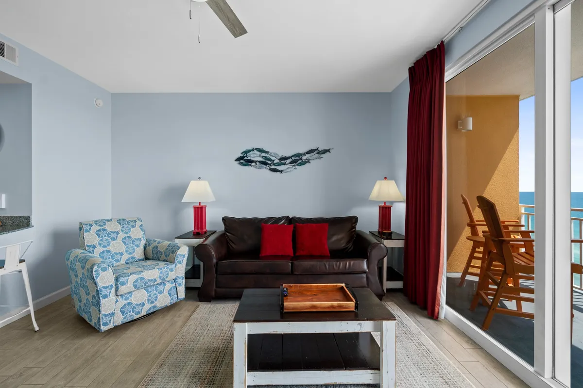 The living room displays a brown leather sofa with red cushions, a blue patterned armchair, and a large sliding glass door leading to a balcony with captivating ocean views. A wooden coffee table sits at the center, and the room’s warm, tropical-inspired decor invites relaxation and socializing.