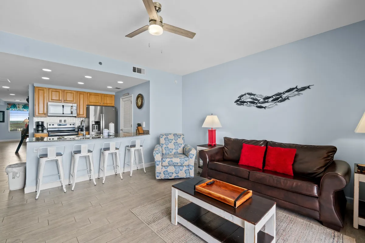 A welcoming open-plan living and kitchen area with a brown leather sofa, red cushions, a kitchen with stainless steel appliances, wooden cabinetry, and a bar counter with white bar stools. The blue patterned armchair and overall decor enhance the inviting, home-like feel.