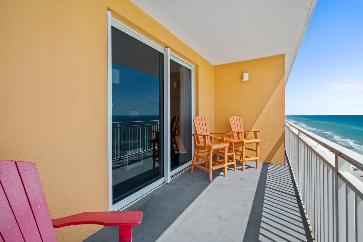 A private condo balcony on a high floor with colorful chairs arranged to face a sweeping ocean view. The coastline stretches into the distance, and the sliding glass doors connect the balcony to the condo's interior. Shadows from the railing create a relaxed, patterned effect on the floor.