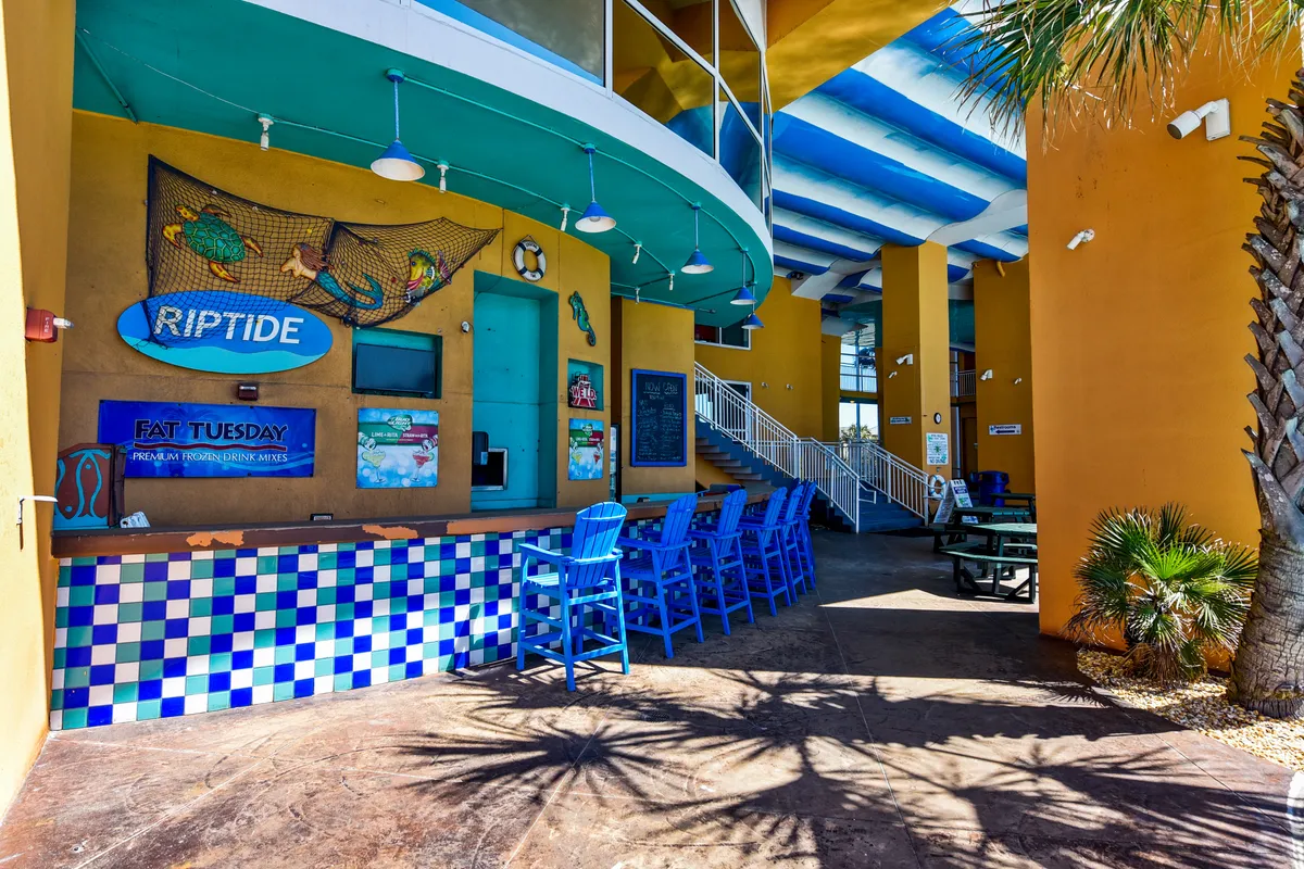 A vibrant, tropical-themed outdoor bar area features a colorful checkered counter, blue bar stools, and playful wall art with tropical fish and netting. Palm tree shadows add to the resort vibe, suggesting a lively yet intimate spot for cocktails and socializing.