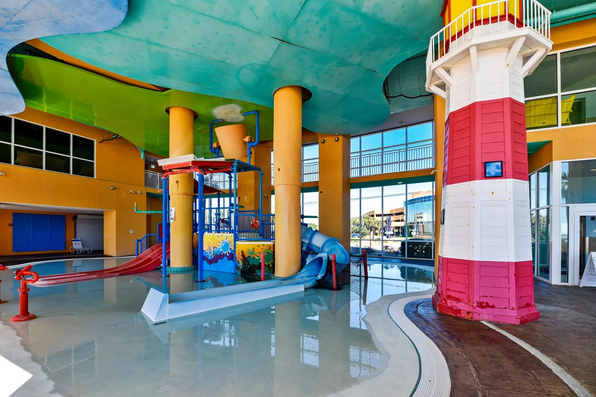 An interior splash zone designed for children, featuring a colorful water play structure with slides, a large red and white lighthouse replica, and a shallow splash pool. The area is filled with bright colors, safe play zones, and families enjoying the space.