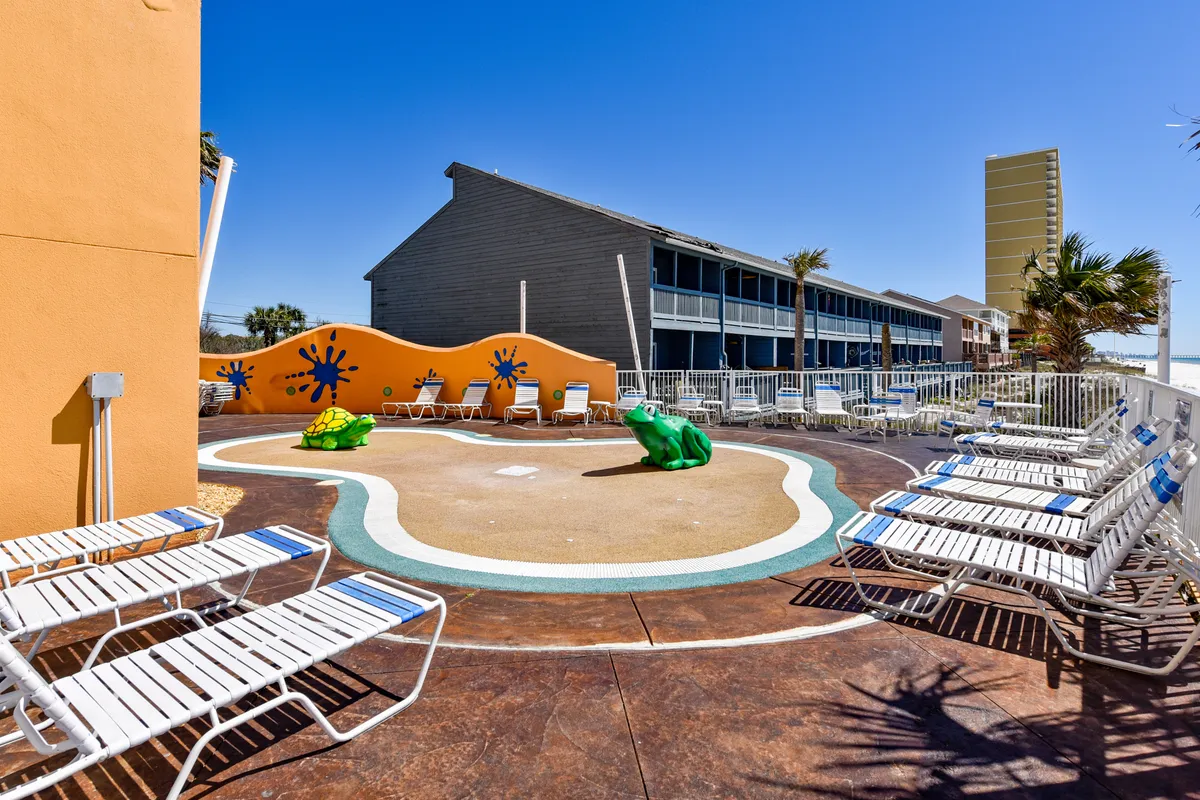 A lively children's splash pad area with playful turtle and frog water sculptures, rows of white lounge chairs for parents, and a multi-story resort building in the background. Kids are seen enjoying the water features while families relax poolside.