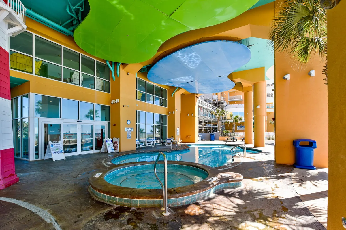 The image features circular hot tubs sheltered by a vibrant overhead canopy, orange structural columns, and glass doors leading indoors. Guests are seen relaxing in the hot tubs and socializing poolside, surrounded by a lively resort atmosphere.