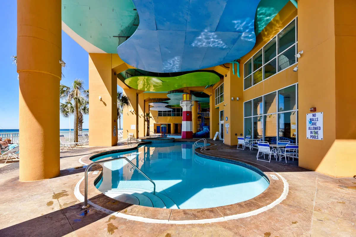 The photo showcases a curved swimming pool with blue and green ceiling panels, tall yellow columns, and a decorative lighthouse structure in the background. Guests can be seen relaxing and playing in the pool, suggesting a family-friendly water park environment.