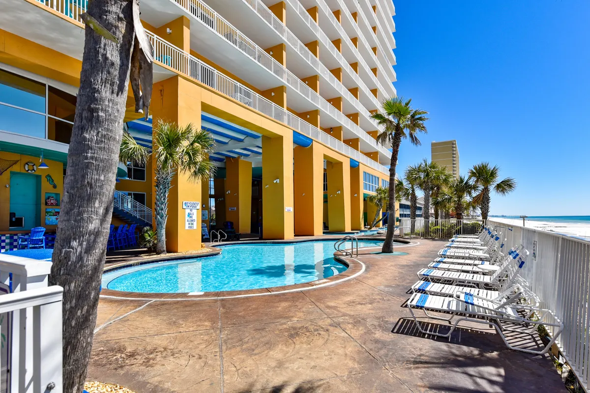 A relaxing Gulf-side heated swimming pool is featured, with rows of white lounge chairs, tall palm trees, and a multi-story yellow and blue resort building in the background. The sparkling pool overlooks the ocean, creating a seamless blend of water recreation and beachside serenity.
