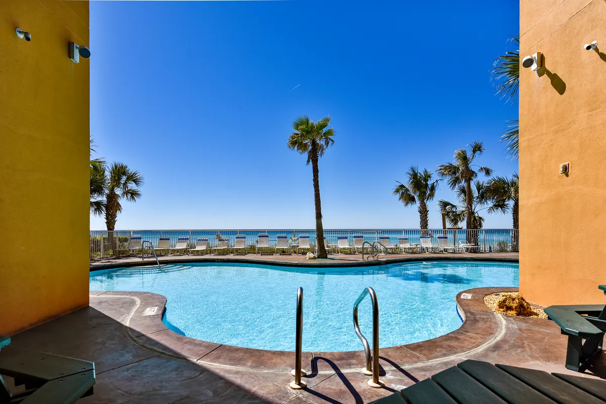A curved swimming pool surrounded by palm trees, with the ocean horizon visible beyond. Lounge chairs are neatly arranged along the fence, inviting guests to sunbathe or relax poolside. The overall setting captures a tropical, resort-style vibe with both pool and ocean amenities in view.