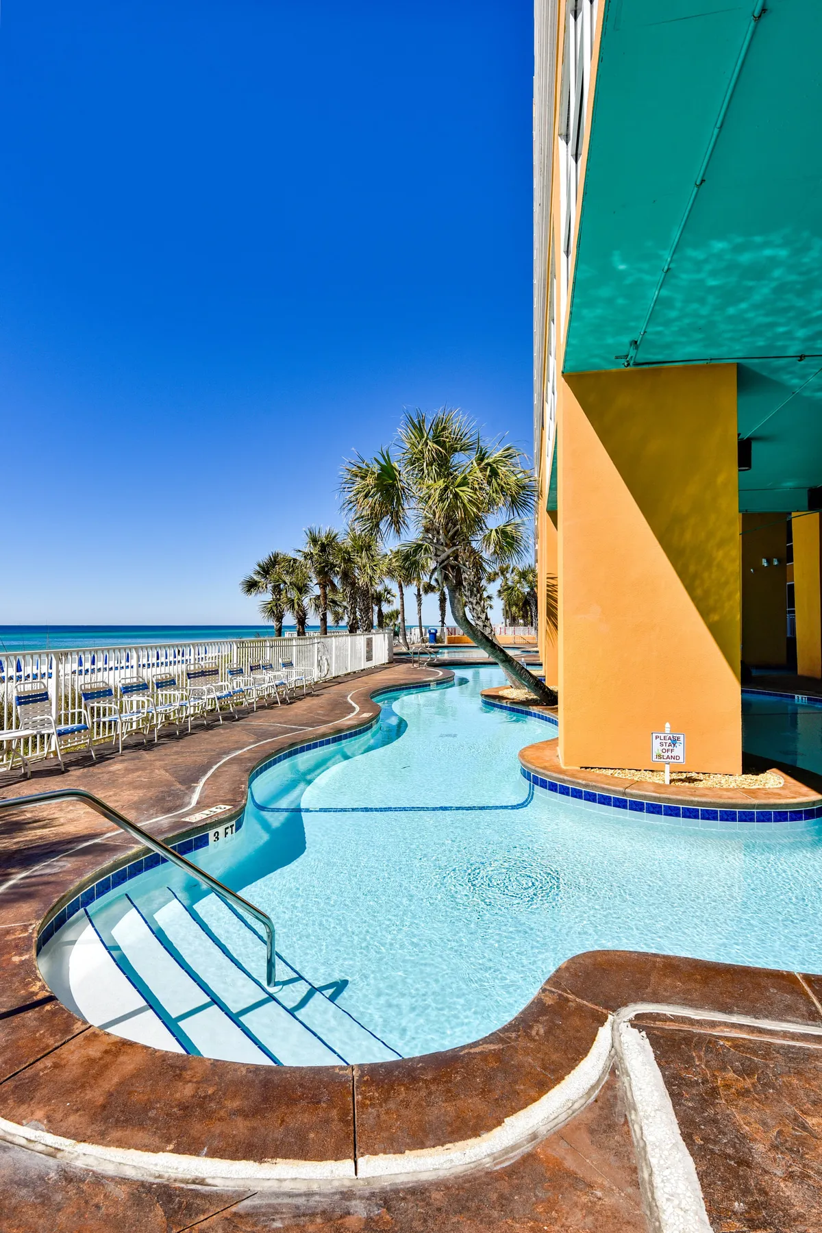 A curved swimming pool lined with palm trees and vibrant resort buildings, with the ocean and sandy beach visible in the background. Guests swim and lounge poolside, enjoying the tropical vibe and scenic views.
