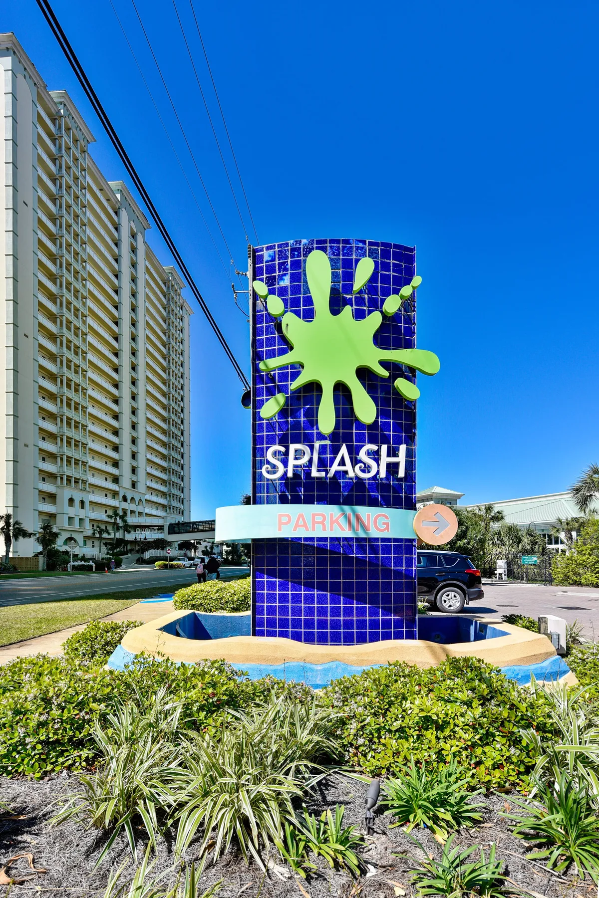 The image features the Splash Resort entrance with its vibrant green splash logo sign, the high-rise resort building in the background, and lush landscaping with green shrubs. Guests are seen arriving and parking, with an inviting atmosphere that hints at nearby water park and beach amenities.