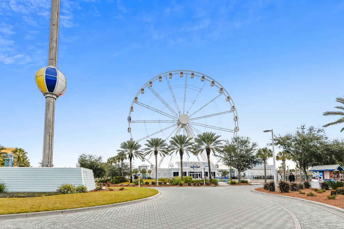 A large Ferris wheel (Sky Wheel) rises above palm-lined walkways and a landscaped plaza, with a curved path inviting leisurely strolls. The cheerful, vacation mood is ideal for couples seeking both adventure and lighthearted fun.