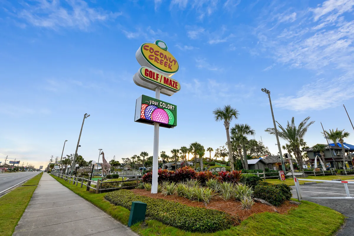 A playful exterior scene showing the Coconut Creek Golf / Maze sign, a whimsical giraffe statue, palm trees, and colorful landscaping. The setting is lively, inviting families and groups for mini golf and maze activities.