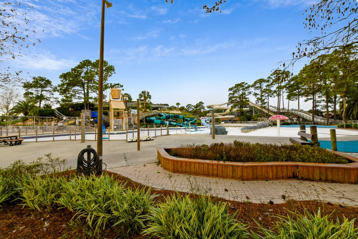 This vibrant exterior image showcases a lively pool area with exciting water slides, a mushroom-shaped water feature, and children playing in the water. Families can be seen enjoying poolside fun, surrounded by lush greenery in wooden planters, all under a bright and inviting sky.