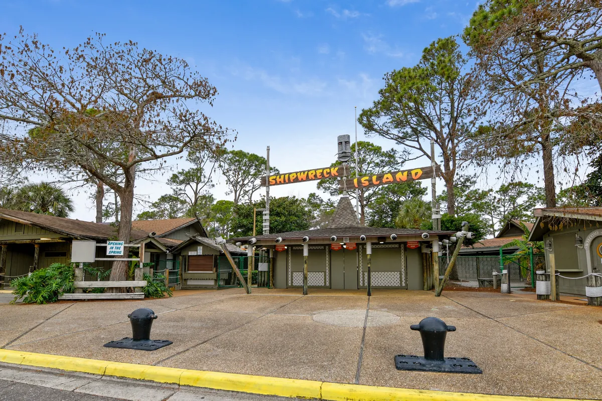 The entrance to Shipwreck Island Water Park, featuring a themed sign, rustic structures, and closed ticket booths, set among trees. The area is calm, suggesting anticipation for the family adventure ahead.